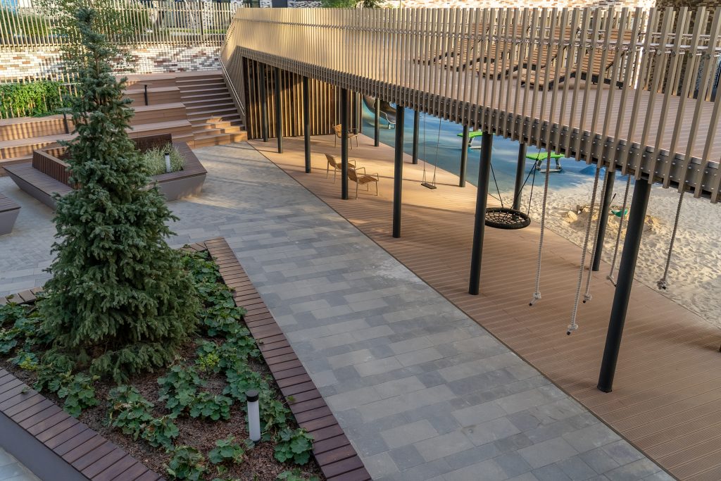 Aménagement d’un espace urbain paysager ou les enfants peuvent profiter des balançoires sous une structure en bois, entourés de verdure luxuriante et de chemins modernes dans cette zone urbaine accueillante